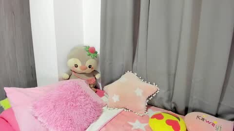 Media: Video of a plush toy rabbit with a red flower, pink fluffy pillow, star pillow, and a yellow pillow with a heart design on a gray bedspread.