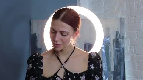 Media: Video of a fair-skinned woman with red hair in a ponytail, wearing a black floral dress with a halter neckline, standing in front of a glowing circular light. Background features a white brick wall and abstract blue painting.