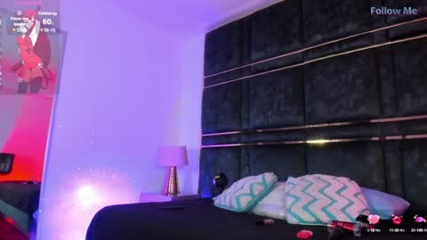 Media: Video of a modern bedroom with a black, textured headboard, teal pillows, and a gold lamp on a white nightstand. A video chat screen displays a red-cloaked figure.