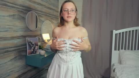 Media: Video of a fair-skinned woman with glasses and shoulder-length blonde hair, wearing a white lace dress, covering her breasts with her hands, standing in a modern bedroom with gray and beige decor.
