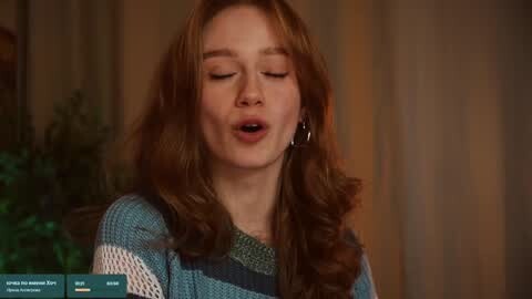 Media: Video of a fair-skinned woman with long, wavy auburn hair, wearing a blue and green striped sweater, singing with eyes closed, background blurred, warm lighting.