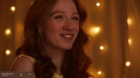 Media: Video of a smiling woman with long, wavy auburn hair, wearing a yellow dress, against a warm, golden background with soft, bokeh lights.