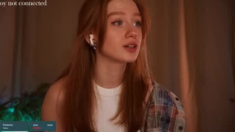 Media: Video of a young, fair-skinned woman with long red hair, wearing a white top, plaid shirt, and white earphones, in a dimly lit room.