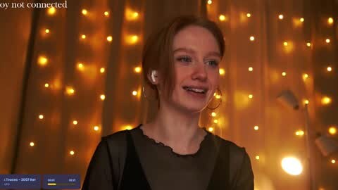 Media: Video of a smiling woman with light skin and brown hair, wearing a black sheer top, in a dimly lit room with warm, golden string lights in the background.