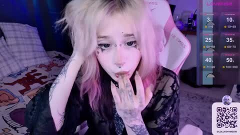 Media: Video of a young woman with platinum blonde hair, black eyeliner, and multiple tattoos, wearing a black lace top, lying on a bed in a dimly lit room.
