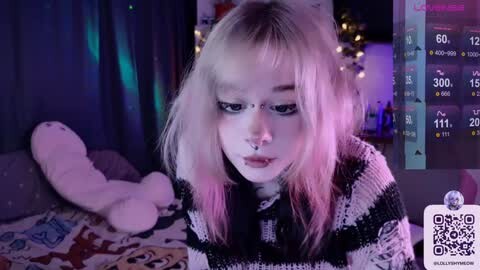 Media: Video of a young woman with platinum blonde hair and dark makeup, wearing a black and white striped sweater, in a dimly lit room with a plush toy, purple lights, and a smartphone screen displaying streaming metrics.