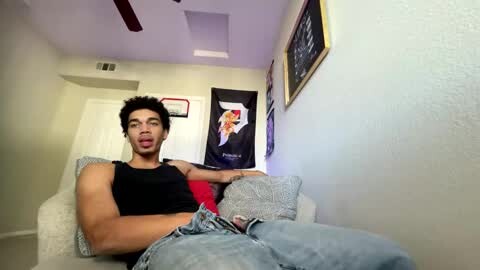 Media: Video of a young man with curly hair lounging on a beige couch, wearing a black tank top and light blue jeans, in a room with light purple walls and framed artwork.