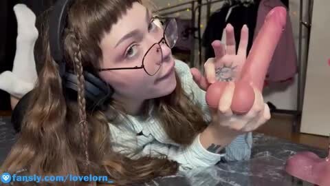 Media: Video of a young woman with glasses, long brown hair, lying on her stomach, holding a large pink dildo, in a bedroom with a bed and clothes in the background.