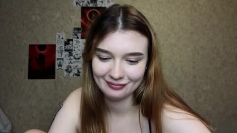 Media: Video of a young woman with long, straight, light brown hair, fair skin, and a small, subtle smile. She wears a black top, and the background features a textured beige wall with black-and-white photos and a red object.