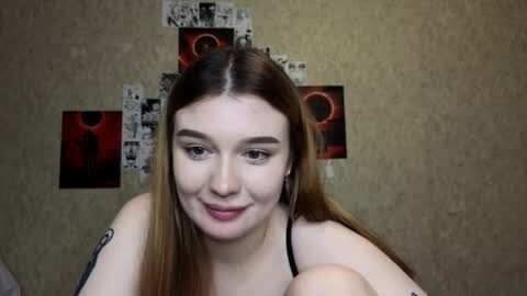 Media: Video of a fair-skinned, light-haired young woman with a tattoo on her upper arm, wearing a black spaghetti strap top, smiling in a dimly lit room with abstract art on the wall.