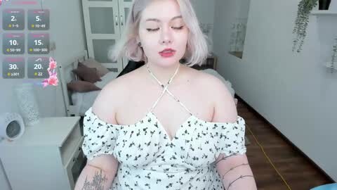 Media: Video of a pale-skinned, plus-sized woman with shoulder-length, platinum blonde hair, wearing a white off-shoulder dress with black star patterns, sitting on a bed in a brightly lit bedroom.