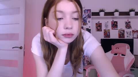 Media: Video of a young woman with light skin, long auburn hair, and a thoughtful expression, resting her chin on her hand. Background features a white wall with framed anime posters and a pink gaming chair.