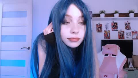 Media: Video of a young woman with pale skin, blue hair, and black sleeveless top, sitting in a gaming chair in a minimalist room with white walls and anime posters.