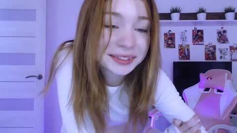 Media: Video of a young woman with fair skin and long, straight brown hair, wearing a white shirt, smiling, in a modern, well-lit room with a white door, shelves with plants, and a gaming chair.