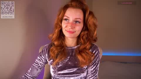 Media: Video of a fair-skinned woman with voluminous, wavy red hair, wearing a velvet top. She has a serene expression and stands against a beige background with purple and blue lighting.