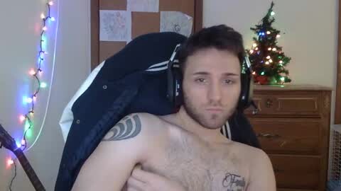 Media: Video of a shirtless man with a beard and headphones, leaning against a wooden dresser with a lit Christmas tree and colorful lights in the background.