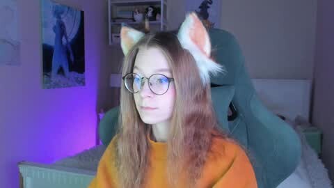 Media: Video of a young woman with long, wavy brown hair, wearing cat ears, glasses, and an orange sweater, seated in a dimly lit, cozy room with a plush chair, bookshelf, and artwork.