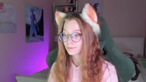Media: Video of a young woman with long, wavy red hair, wearing cat ears and glasses, seated in a room with purple lighting, cat plush toys, and anime posters on the wall.