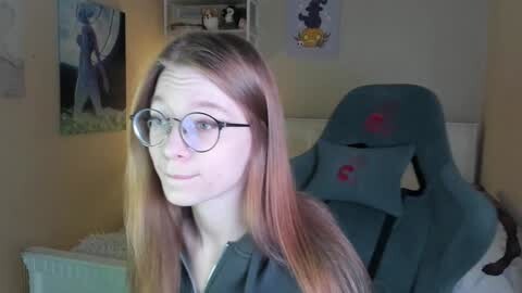 Media: A video of a young woman with long, straight, light brown hair and round glasses, standing in a bedroom with a green gaming chair and posters on the wall.