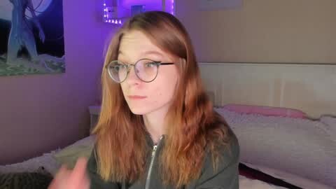 Media: Video of a fair-skinned, red-haired woman with glasses, wearing a green hoodie, sitting on a bed with white pillows and a beige headboard in a dimly lit room with purple lights.