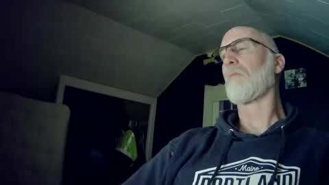 Media: Video of a bald, bearded man in glasses, wearing a black \"Fort Lauderdale\" hoodie, standing in a dimly lit room with dark walls and a partially visible doorway.