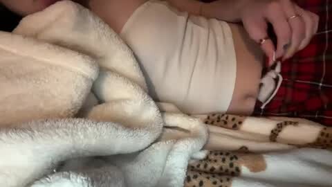 Media: Video of a person's torso covered in a beige top, lying on a bed with a white blanket and a leopard-print blanket, and a red plaid shirt in the background.