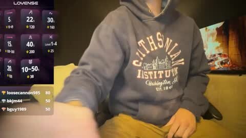 Media: A video of a person in a dark grey \"Gothamist\" hoodie, sitting on a yellow couch, holding a remote, with a TV screen showing a sports game in the background.