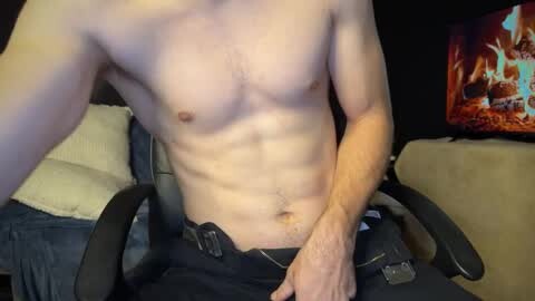 Media: Video of a shirtless, muscular white man with fair skin, wearing black pants, sitting in a gaming chair. Background includes a TV with flames, beige couch, and dark walls.
