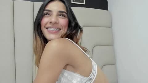 Media: Video of a smiling young woman with straight black hair and braces, wearing a white lace bra. She stands in a white cushioned room with a gray wall.