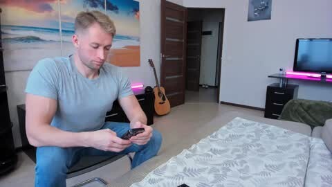 Media: Video of a muscular, blonde man in a light blue shirt, sitting on a bed, holding a smartphone, in a modern bedroom with a guitar, a beach painting, and a flat-screen TV.