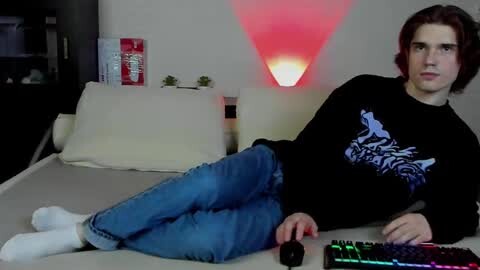 Media: Video of a young white man with shoulder-length brown hair, wearing a black shirt and blue jeans, reclining on a beige bed with a computer mouse on the floor.
