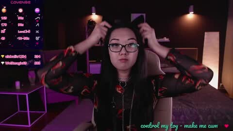 Media: Video of a young Asian woman with long black hair, glasses, and a black floral dress, adjusting her hair. She sits in a dimly lit room with purple lighting, a TV, and a bed visible in the background. Text overlay reads, \"control my boy make me cum.\