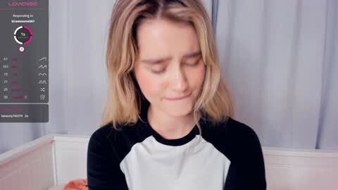 Media: A video of a young woman with shoulder-length blonde hair, wearing a black and white shirt, sitting in a room with light grey curtains.