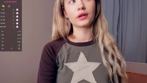 Media: Video of a young, light-skinned woman with long, wavy blonde hair, wearing a grey and black sweatshirt with a large white star. Background shows a simple room with grey walls, a window, and a calendar.