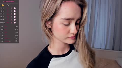 Media: Video of a young, fair-skinned woman with long, blonde hair, wearing a black and white shirt, standing indoors near a window with white curtains.