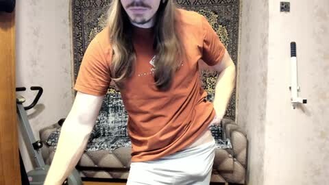 Media: Video of a man with long brown hair, wearing an orange T-shirt and gray shorts, bending slightly, standing in a room with a patterned tapestry and a beige couch in the background.