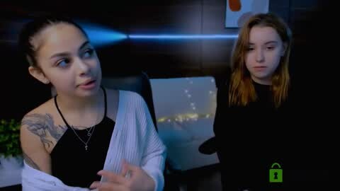 Media: Video of two young women, one with dark hair and tattoos, speaking in a dimly-lit room with a cityscape painting on the wall.