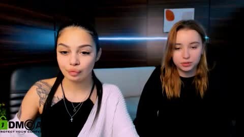 Media: Video of two young women with different skin tones and hair textures sitting side by side in a dimly lit room. One wears a black tank top and a pink cardigan, while the other is in a black sweater.