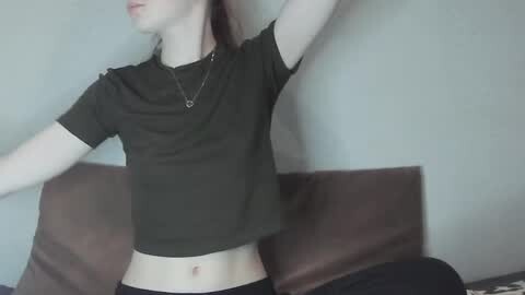 Media: Video of a slender, pale-skinned woman with red hair, wearing a dark T-shirt and black pants, stretching against a beige wall.