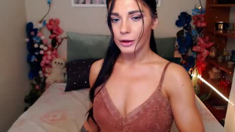 Media: Video of a young woman with long black hair, wearing a pink lace bra, standing in a cozy bedroom adorned with floral decor and stuffed toys.