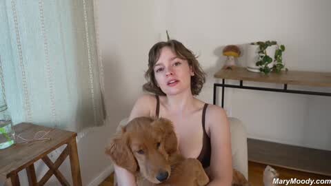 Media: Video of a fair-skinned woman with short brown hair, wearing a brown tank top, sitting on a white chair holding a golden retriever puppy. The background features a wooden table with a vase and green plants, beige curtains, and a wooden floor.