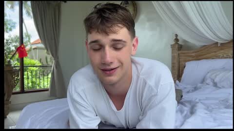 Media: Video of a young, fair-skinned, short-haired Caucasian man with closed eyes and a relaxed smile, wearing a white T-shirt, lying on a bed with white sheets in a bright, minimalist room with a wooden headboard and large window.