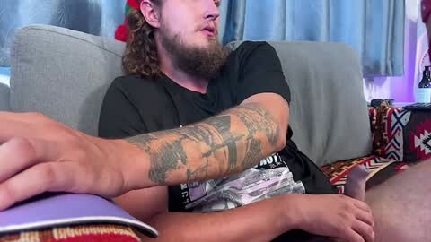 Media: Video of a bearded man with a tattooed arm, wearing a black t-shirt, reading a purple book on a gray couch, in a living room with patterned cushions and curtains.