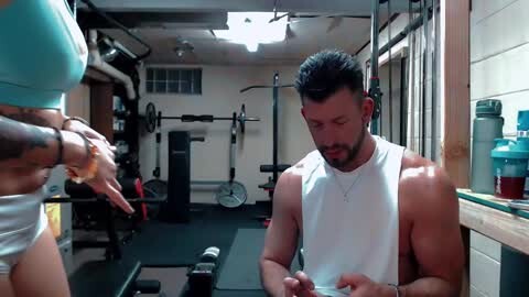 Media: Video of a muscular, bearded man in a white tank top, concentrating on his phone in a well-equipped, dimly-lit gym with a woman in a teal top in the background.