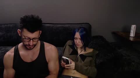 Media: A video of a muscular, black-haired man in a black tank top and glasses, sitting on a dark couch with a blue-haired woman in an olive jacket, both focused on smartphones.