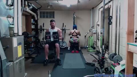 Media: Video of a dimly lit home gym with a muscular man and a slender woman exercising on separate machines. The room is cluttered with equipment and has a beige wall and exposed pipes.