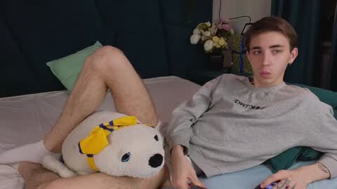 Media: Video of a young man in a gray sweater lying on a bed with a plush dog, wearing only socks, in a dark room with green pillows and a floral arrangement.