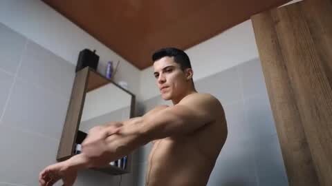 Media: Video of a muscular, shirtless young man with short dark hair stretching in a modern, beige-tiled bathroom with a wooden door and mirror.