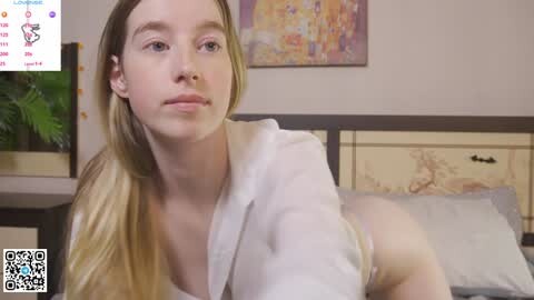 Video of a young woman with long blonde hair, wearing a white shirt, sitting on a bed in a bedroom with colorful abstract art on the wall and a potted plant.