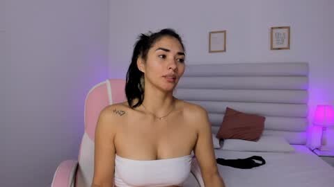 Media: Video of a Latina woman with long black hair, medium skin tone, wearing a white strapless dress, sitting on a pink chair in a modern bedroom with a white headboard and framed art on the wall.